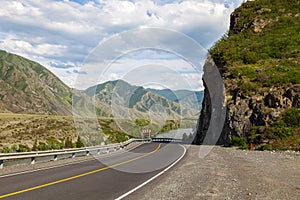 A new winding asphalt black road with a yellow dividing strip between the rocks and the Katun River in the Altai mountains under a