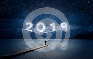 New year background, young man standing on a jetty in a lake and looking to the mountains under the dark sky with cloudy text 2019