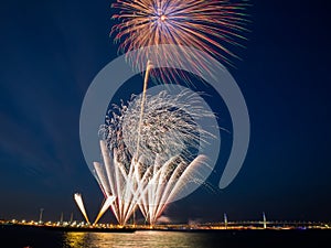 Bright fireworks burst in the blue sky with distant city lights and sea water