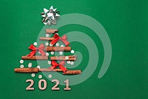 New year card 2021. Christmas tree made of cinnamon sticks with silver gift bow and small red bows on green background with silver