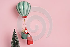 New Year card. toy Santa Claus is flying on the balloon near Christmas tree and holding red paper bag. pink background with copy