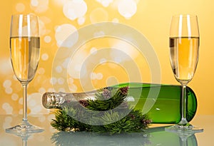 New year champagne bottle lies on the table near pine branches with pine cones and two full wine glass, on yellow background