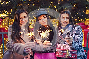 New Year concept. Women friends burning sparklers in Lviv by Christmas tree. Girls holding shopping bags and gifts