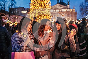 New Year concept. Women friends burning sparklers in Lviv by Christmas tree on street fair. Girls holding shopping bags