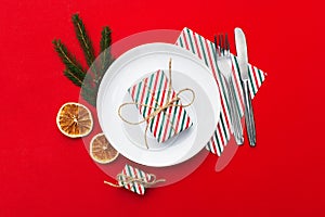 New year dinner table place setting with plate, present box, christmas eve fir branch on red background. Minimal frame