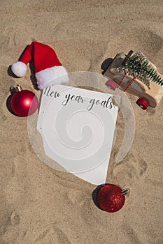 NEW YEAR GOALS text on paper greeting card on background sandy beach sun coast. Christmas balls Santa hat New Year New