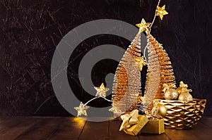 New Year home decor in dark and golden color - christmas trees, glowing lights, stars, balls, gift box, ribbons in black interior.
