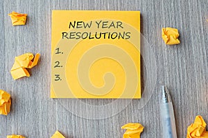 NEW YEAR RESOLUTIONS word on yellow note with  pen and crumbled paper on wooden table background. New start, Strategy and Goal