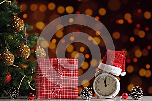 New Year`s composition. Gift box and alarm clock in a Santa hat next to a decorated Christmas tree on a blurred background of