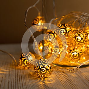 New Year`s garland. Golden metal balls in glass jar on light wooden table background. Christmas yellow gold garland lights from