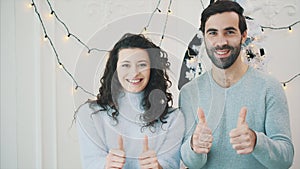 New year video of childish couple near christmas tree, giving thumbs up and smiling.