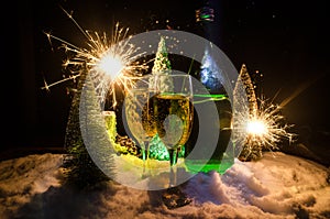 New Years Eve celebration background with pair of flutes and bottle of champagne with christmas tree on snow on dark background.