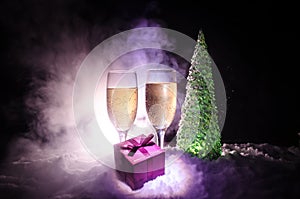 New Years Eve celebration background with pair of flutes and bottle of champagne with christmas tree on snow on dark background.