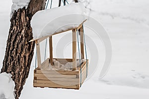 A new yellow bird and squirrel feeder house from plywood with white snow on the roof is hanging on a red rope on a brown