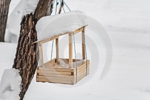 A new yellow bird and squirrel feeder house from plywood with white snow on the roof is hanging on a red rope on a brown