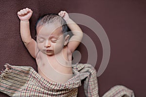 Newborn baby boy sleeping peacefully on brown blanket with both hands up posture. checkered cloth on brown background.new born