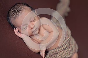 newborn baby boy sleeping peacefully on brown blanket with checkered cloth on brown background.new born portrait