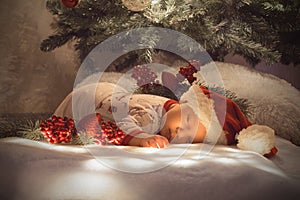 Newborn baby boy sleeping under Christmas tree near lot of decorations. Wearing SantaÃÂ´s hat