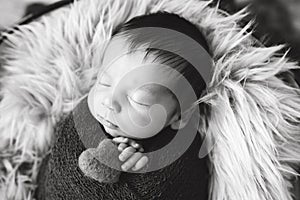 Newborn baby boy wrapped in brown wrap on furry blanket. Baby sleeping. Infant with long dark hair. The baby is holding a heart in