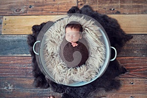 Newborn baby boy wrapped in brown wrap on furry blanket in pot on wooden vintage floor . Baby sleeping. Infant with long dark hair