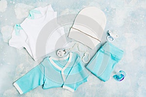 Newborn baby clothes - white bodysuit, blue shirt, blue pants, white hat and pacifier on blue textured background. Baby shower