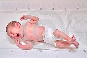 A newborn baby is crying on a white changing table with a ruler for measuring growth