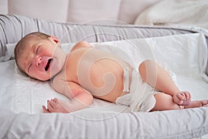 A newborn baby in a diaper is crying on the bed. Child is a boy in a cocoon on a cot