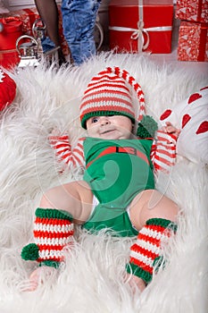 Newborn baby girl dressed in gnome costume lying on white fur carpet among christmas decorations.