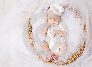 Newborn Baby Girl in White Headband Bow sucking Pacifier sleeping in Basket over White Tulle Fabric. One Month Child dreaming in