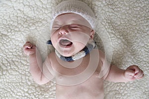 A newborn baby in hat and scarf is crying on a light background