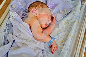 A newborn baby with a maternity hospital bracelet on his arm is sleeping in a crib. A newly born child in a clinic bed behind a