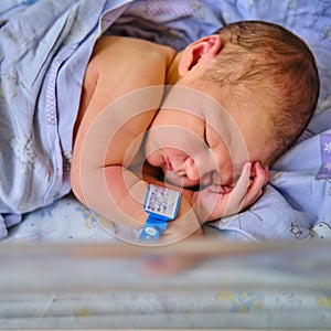 A newborn baby with a maternity hospital bracelet on his arm is sleeping in a crib. A newly born child in a clinic bed behind a