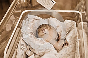 A newborn baby with a maternity hospital bracelet on his arm is sleeping in a crib. A newly born child in a clinic bed behind a