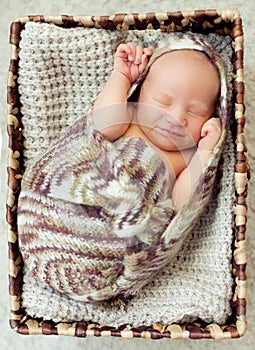 A newborn baby sleeps in a drawer wrapped in a knitted brown scarf on the white carpet