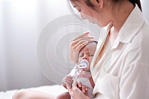 Newborn baby with teat was held by her Asian mother who is sitting on the bed and look to baby face with love