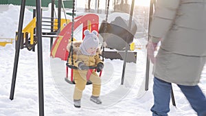 A newborn baby in yellow winter clothes swings on a swing for toddlers during a snowfall on a winter playground