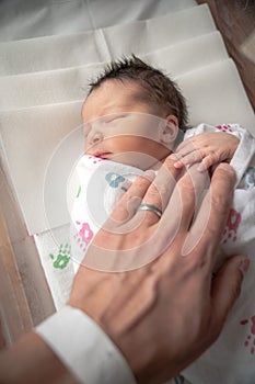 A newborn infant baby girl in a blanket swaddle rests her tiny hand and fingers on her fathers wrinkled hand as she sleeps