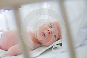 The newborn is lying in a crib with white rods. The child looks through the bars of the white bed. Meaningful look of a newborn. A