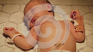 Newborn Is Lying Under Warm Lamp On Changing Table With Name Pendants On His Arm