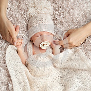 Newborn sucking Pacifier in Sleep. Parent holding Baby Fist in Hands. One Month little Child with Dummy in knitted Hat covered