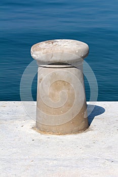 Newly made dark grey concrete mooring bollard looking like mushroom on top of concrete pier with calm clear blue sea in background