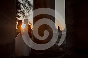 Newlywed couple in love is tenderly hugging among old columns during the bright sunset.