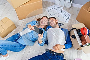 Newlywed couple lying down on the wooden floor of their new apartment and making selfie by smart phone. Top view.