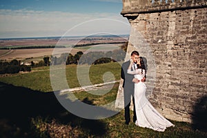 Newlywed couple posing near old castle wall, fairytale wedding at ancient castle outdoors, bride and groom hugging near fortress