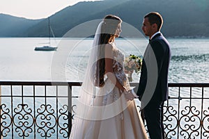 Newlyweds couple, bride and groom holding hands looked into each other`s eyes on a lake background. Cute girl in white