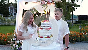 Newlyweds cut wedding cake on warm summer sunset near decorated arch outdoors. Bride and groom smiling and cutting