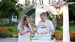 Newlyweds cut wedding cake on warm summer sunset near decorated arch outdoors. Bride and groom smiling and cutting
