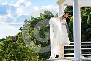 The newlyweds happily embrace in a beautiful gazebo with columns against the backdrop of foliage and sky