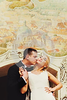 Newlyweds kiss standing behind a wall with city fresco