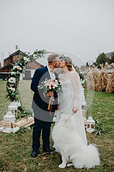 Newlyweds stand near wedding arch and kiss next to dog